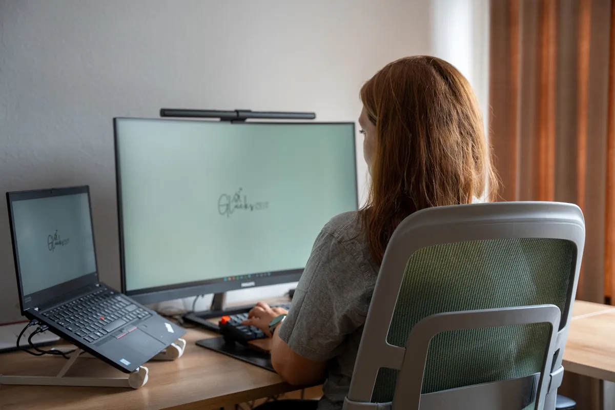 Mitarbeiterin sitzt im Büro am Schreibtisch vor Bildschirm und Laptop mit dem Glückszeit-Logo