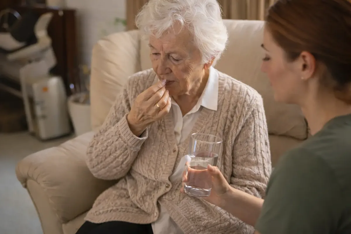 Seniorin im Sessel nimmt eine Tablette, eine Pflegerin reicht ihr ein Glas Wasser